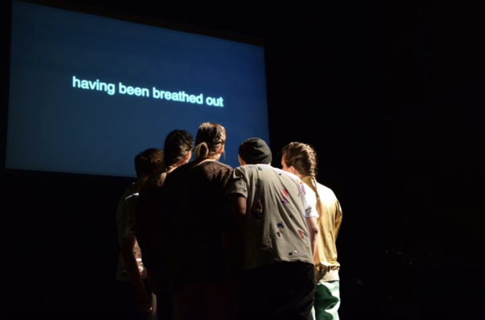 A group of peoples huddles inwards, standing infront of a screen with pale blue text on a dark blue background that reads "having been breathed out"