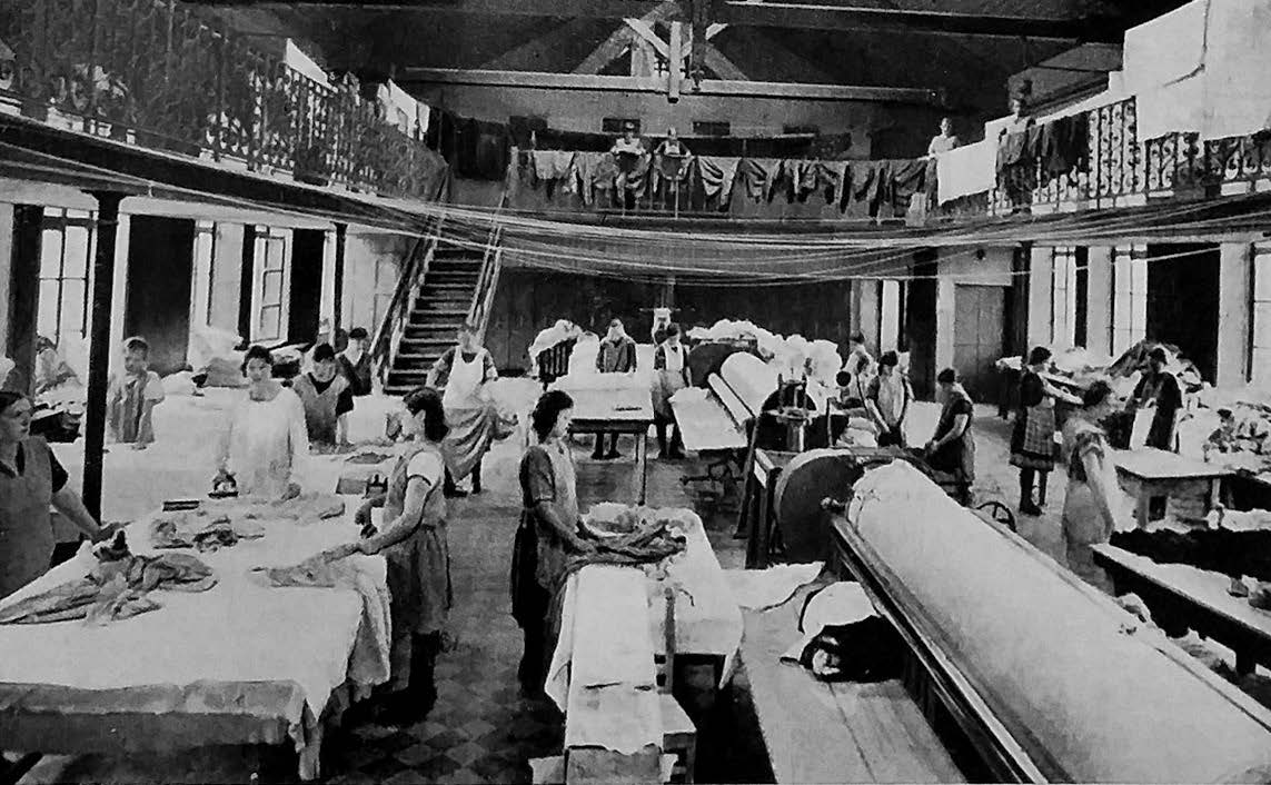 a laundry room - a large high ceiling room with many women working at tables with large machinery and cloth