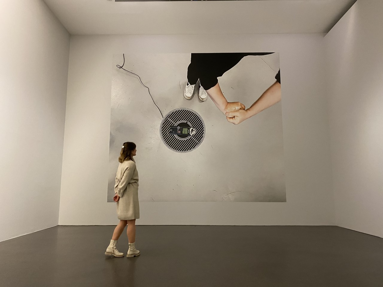 The artwork - a large photograph of Salomé's hands using Nivea cream over a zoom recorder on the floor. The artwork is installed as a large photograph filling a gallery wall, a person stands infront