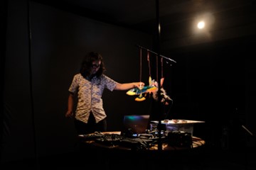 Kate Carr touches a crafted bird hanging alongside a series of homemade objects. Below is a laptop and wires. She stands in a darkened performance space