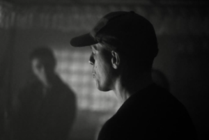 Luciano wearing a cap looks away from the camera in black and white. Blurred figure in background